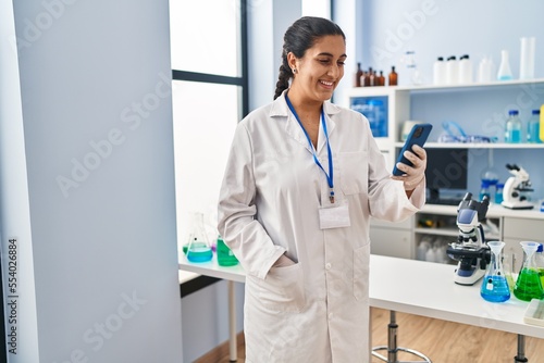 Young hispanic woman wearing scientist uniform using smartphone at laboratory