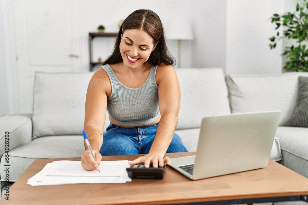 Young beautiful hispanic woman writing on document using calculator at home Young beautiful hispanic woman writing on document using calculator at home