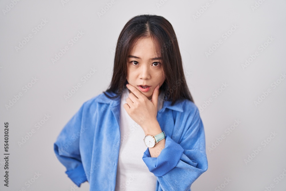 Young chinese woman standing over white background looking fascinated with disbelief, surprise and amazed expression with hands on chin Young chinese woman standing over white background looking fascinated with disbelief, surprise and amazed expression with hands on chin