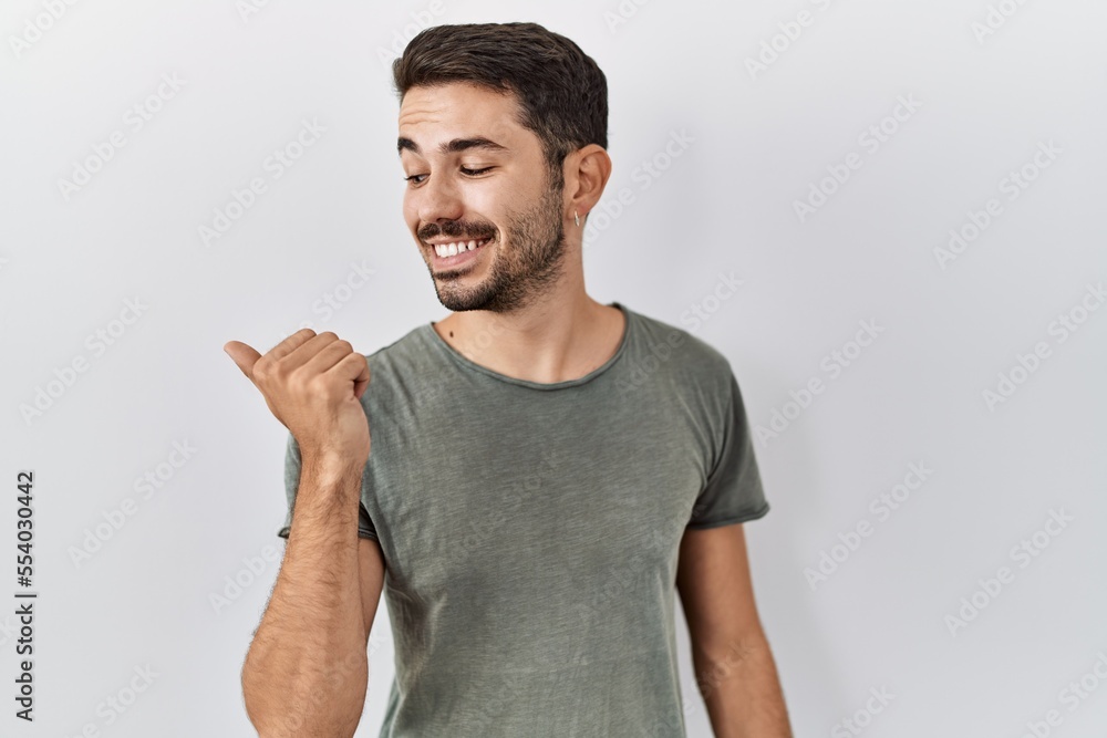 Young hispanic man with beard wearing casual t shirt over white background smiling with happy face looking and pointing to the side with thumb up. Young hispanic man with beard wearing casual t shirt over white background smiling with happy face looking and pointing to the side with thumb up.
