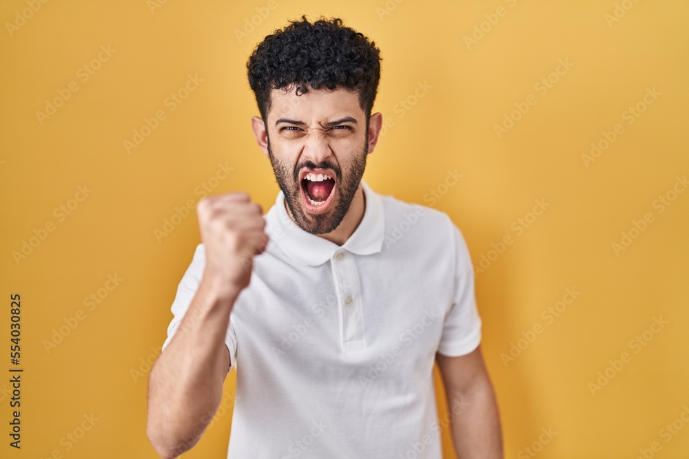 Arab man standing over yellow background angry and mad raising fist frustrated and furious while shouting with anger. rage and aggressive concept. Arab man standing over yellow background angry and mad raising fist frustrated and furious while shouting with anger. rage and aggressive concept.
