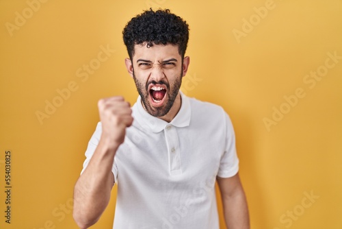 Arab man standing over yellow background angry and mad raising fist frustrated and furious while shouting with anger. rage and aggressive concept.