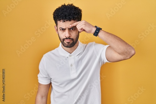 Arab man standing over yellow background pointing unhappy to pimple on forehead  ugly infection of blackhead. acne and skin problem