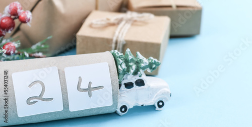 Toy car with Christmas tree, toilet paper tube with number 24 and craft gifts with fir branches on a blue background.