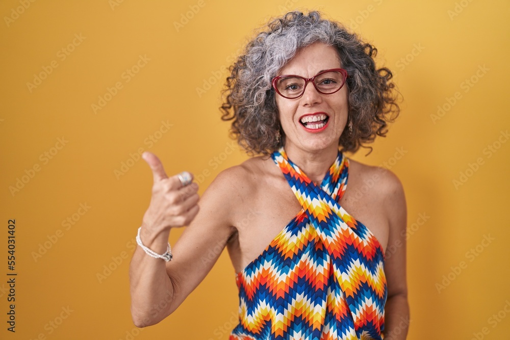 Middle age woman with grey hair standing over yellow background doing happy thumbs up gesture with hand. approving expression looking at the camera showing success.