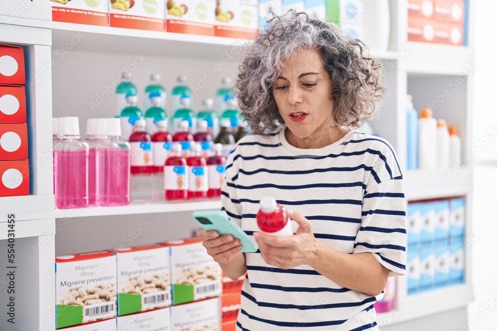 Middle age grey-haired woman client using smartphone holding medication bottle at pharmacy Middle age grey-haired woman client using smartphone holding medication bottle at pharmacy