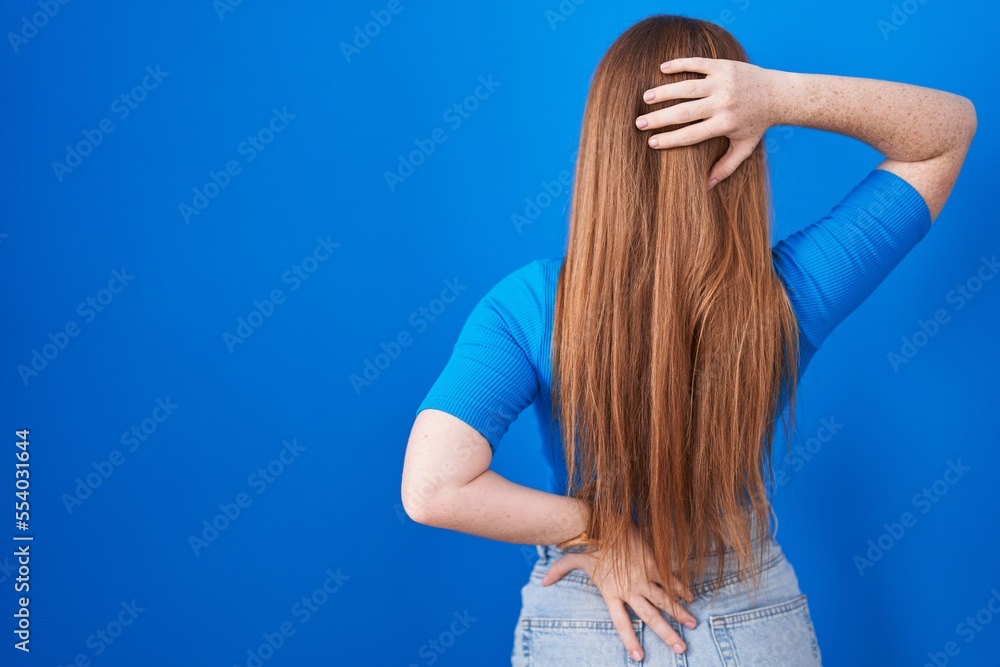 Redhead woman standing over blue background backwards thinking about doubt with hand on head Redhead woman standing over blue background backwards thinking about doubt with hand on head