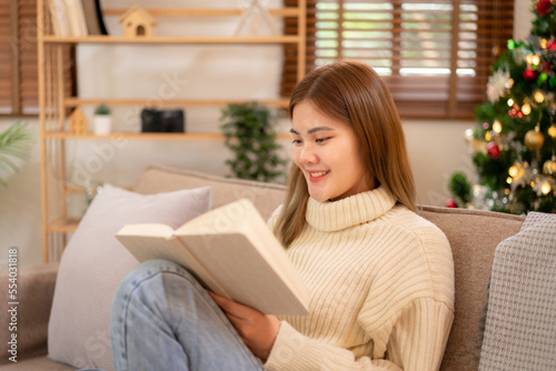 Young asian woman wearing sweater and reading a book while sitting to relaxation in holiday on sofa in cozy