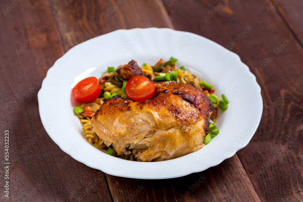 Pasta with vegetables and baked chicken leg sprinkled with green onions in a plate on a wooden table.