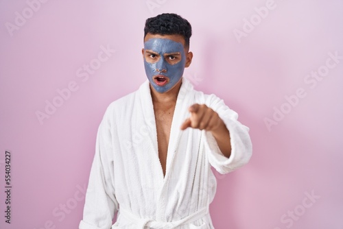 Young hispanic man wearing beauty face mask and bath robe pointing displeased and frustrated to the camera, angry and furious with you