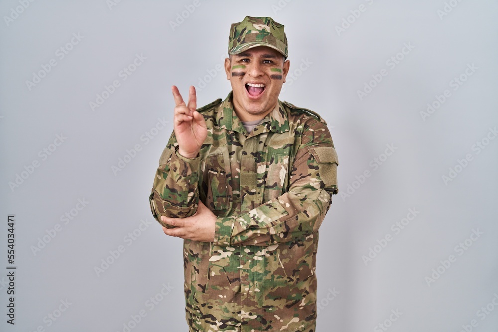Hispanic young man wearing camouflage army uniform smiling with happy face winking at the camera doing victory sign. number two. Hispanic young man wearing camouflage army uniform smiling with happy face winking at the camera doing victory sign. number two.