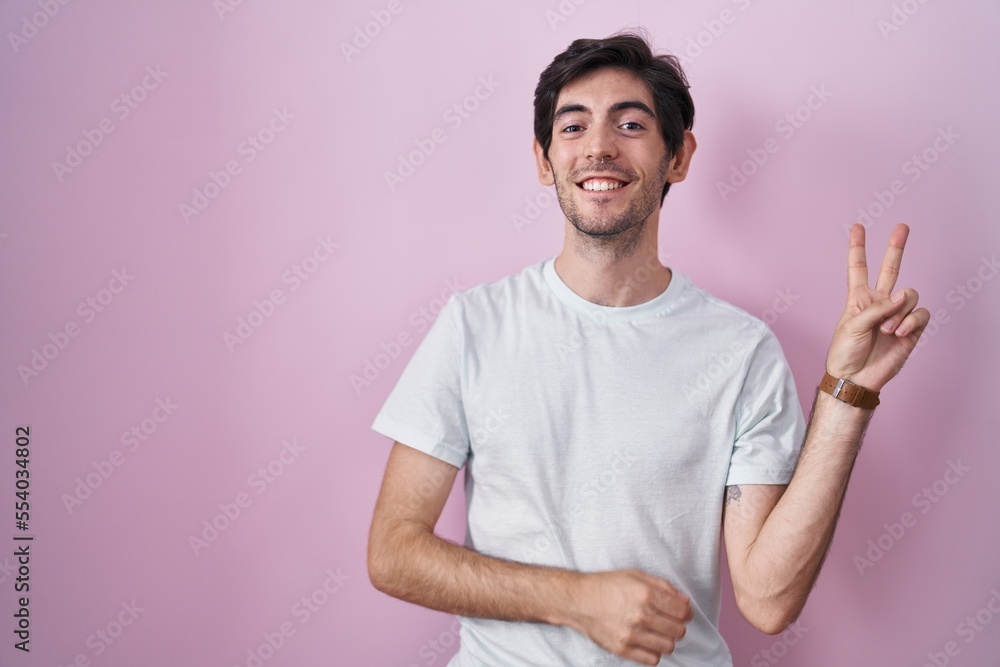 Obraz premium Young hispanic man standing over pink background smiling with happy face winking at the camera doing victory sign with fingers. number two.