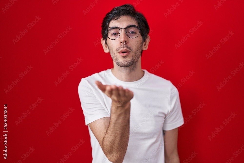 Young hispanic man standing over red background looking at the camera blowing a kiss with hand on air being lovely and sexy. love expression. Young hispanic man standing over red background looking at the camera blowing a kiss with hand on air being lovely and sexy. love expression.