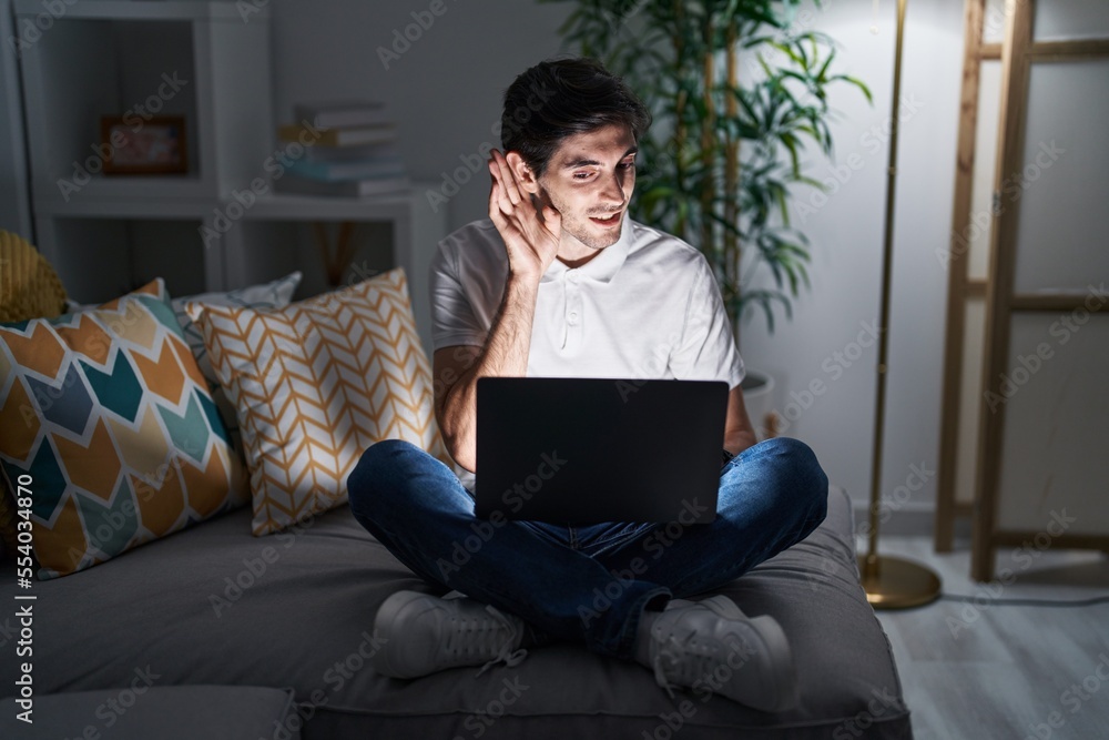 Young hispanic man using laptop at home at night smiling with hand over ear listening an hearing to rumor or gossip. deafness concept. Young hispanic man using laptop at home at night smiling with hand over ear listening an hearing to rumor or gossip. deafness concept.