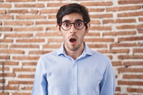 Young hispanic man standing over brick wall background afraid and shocked with surprise and amazed expression, fear and excited face. © Krakenimages.com