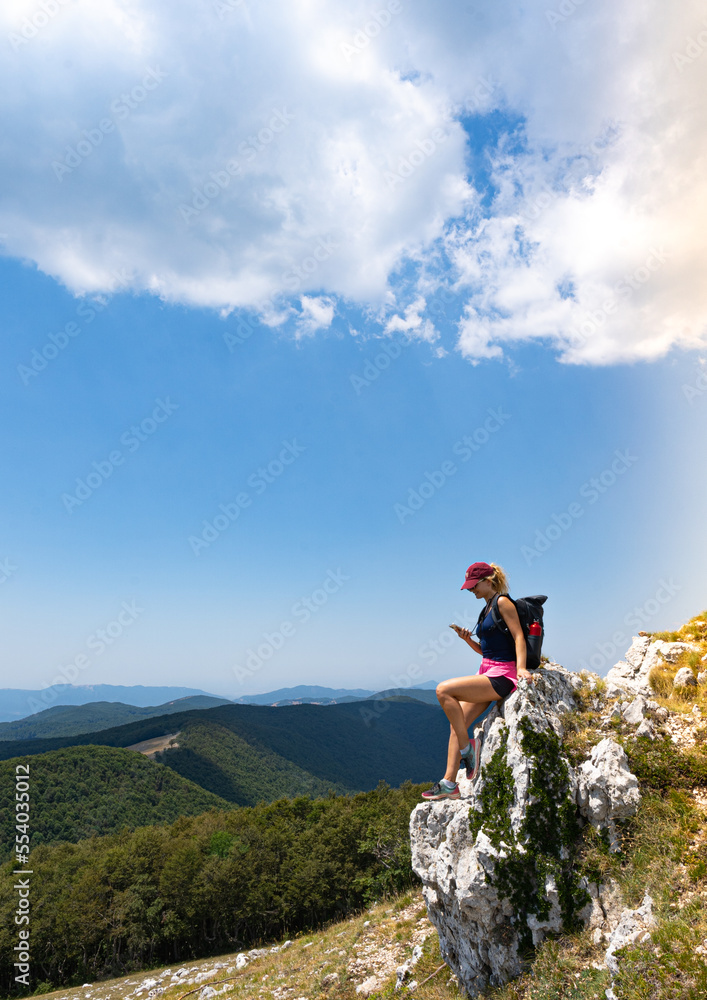 vertical photo of explorer woman sitting while watching the mobile phone in a panoramic view of mountains
