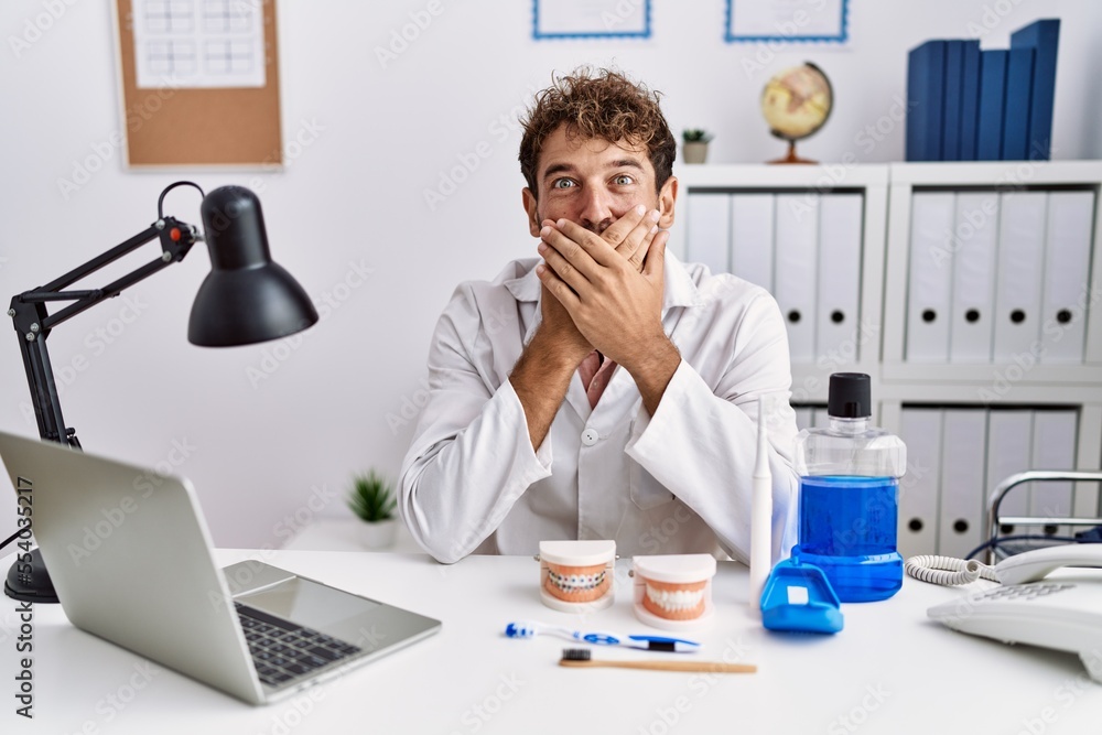 Young hispanic dentist man working at medical clinic shocked covering mouth with hands for mistake. secret concept. Young hispanic dentist man working at medical clinic shocked covering mouth with hands for mistake. secret concept.