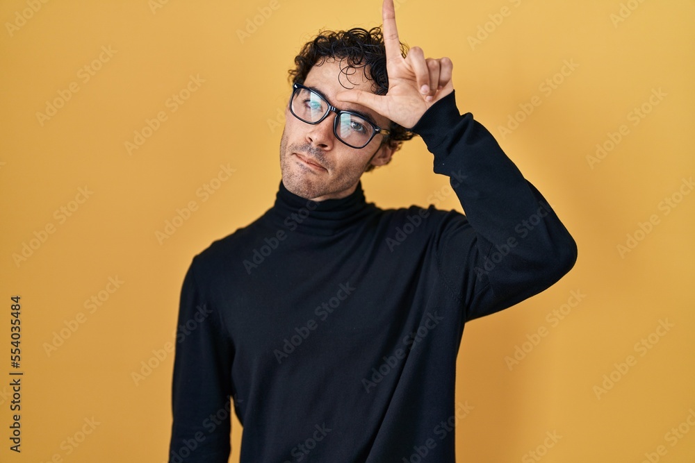 Hispanic man standing over yellow background making fun of people with fingers on forehead doing loser gesture mocking and insulting. Hispanic man standing over yellow background making fun of people with fingers on forehead doing loser gesture mocking and insulting.