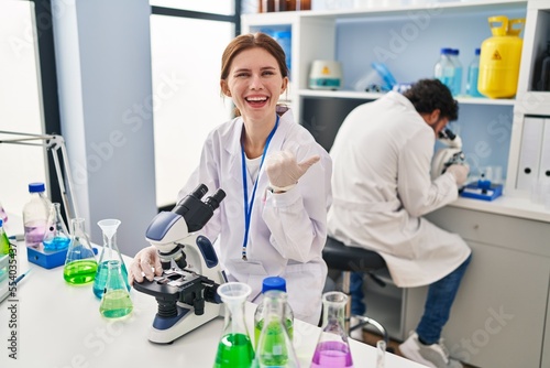 Young two people working at scientist laboratory pointing thumb up to the side smiling happy with open mouth