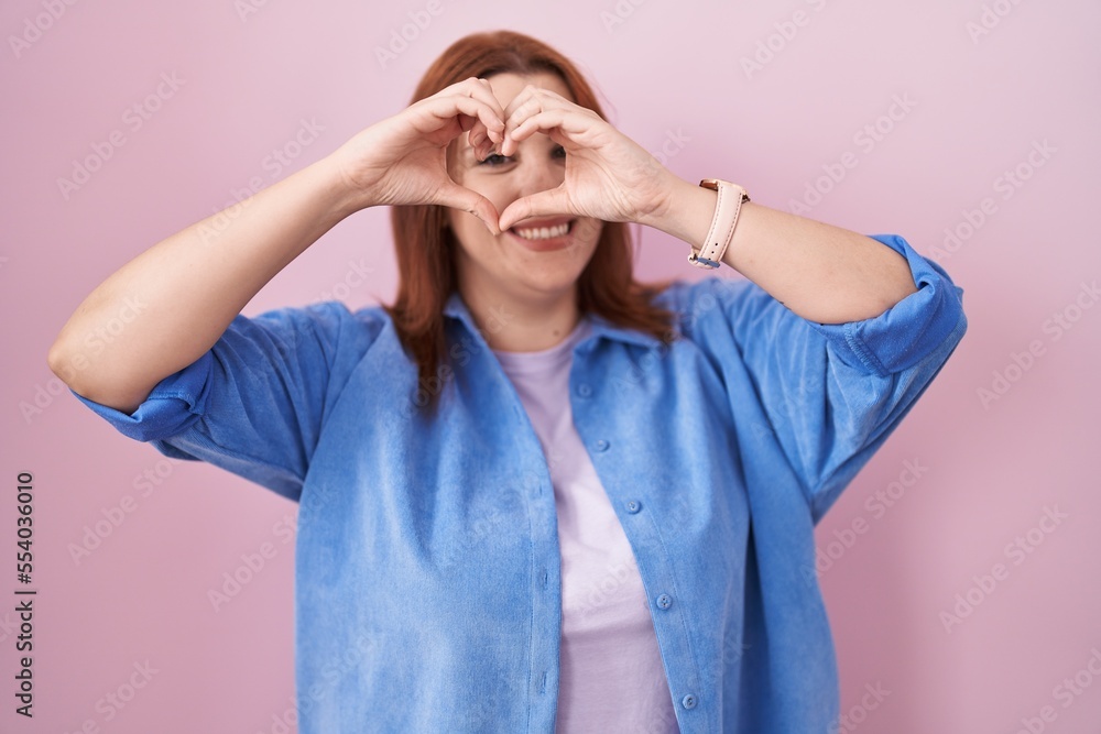 Young hispanic woman with red hair standing over pink background doing heart shape with hand and fingers smiling looking through sign Young hispanic woman with red hair standing over pink background doing heart shape with hand and fingers smiling looking through sign