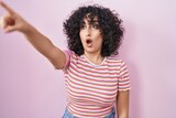 Young middle east woman standing over pink background pointing with finger surprised ahead, open mouth amazed expression, something on the front