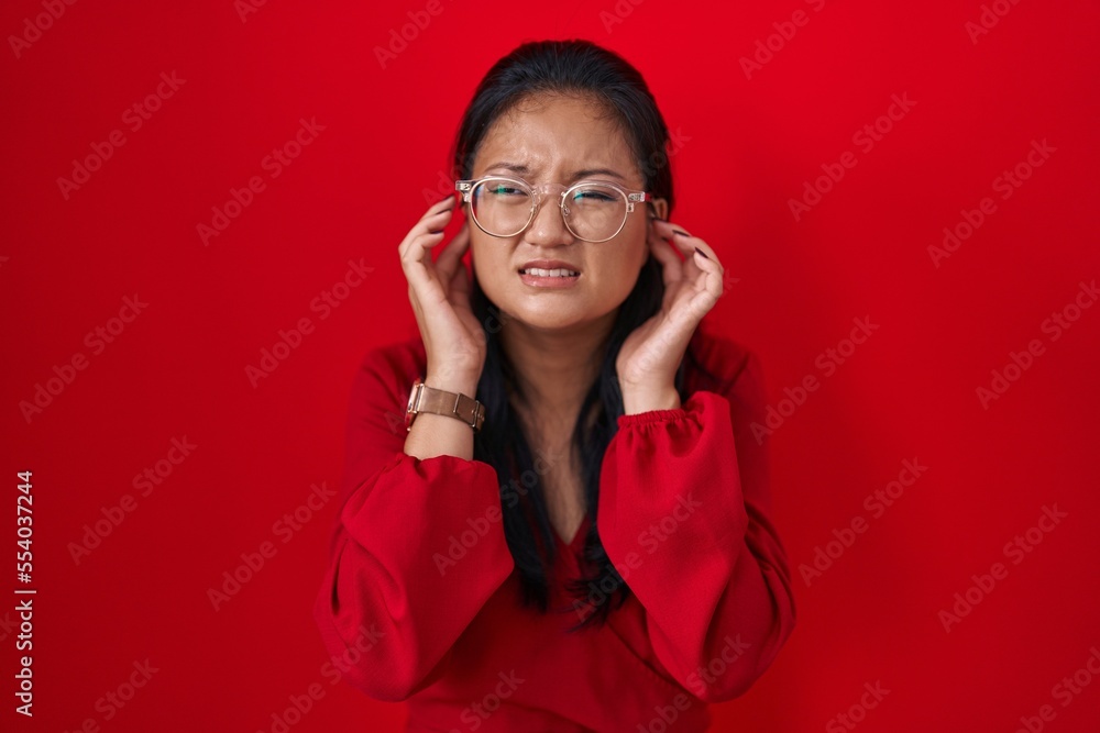 Asian young woman standing over red background covering ears with fingers with annoyed expression for the noise of loud music. deaf concept. Asian young woman standing over red background covering ears with fingers with annoyed expression for the noise of loud music. deaf concept.