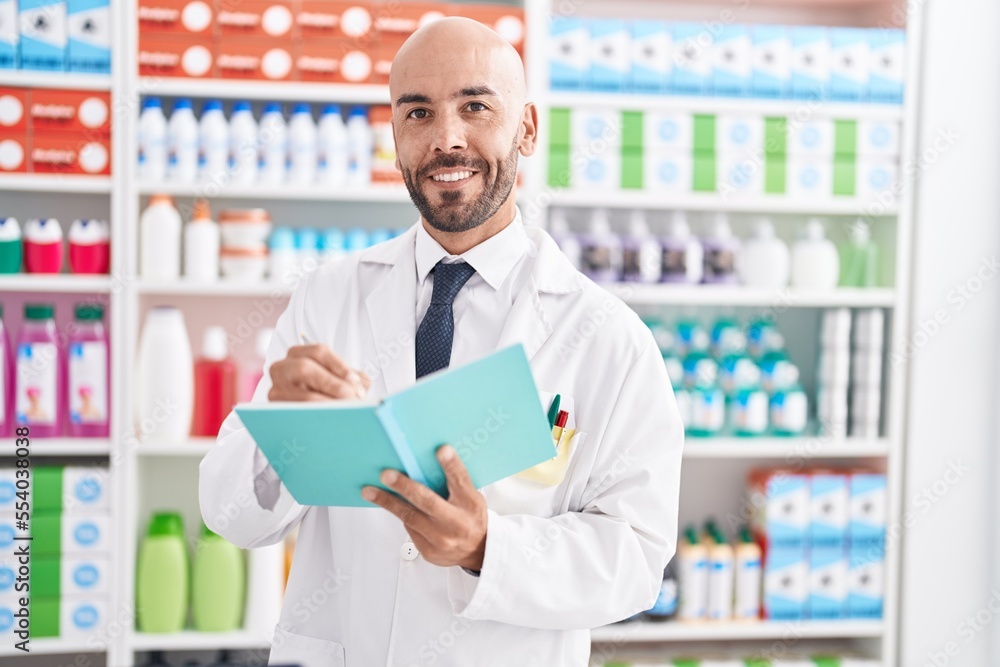 Young bald man pharmacist smiling confident writing on document at pharmacy