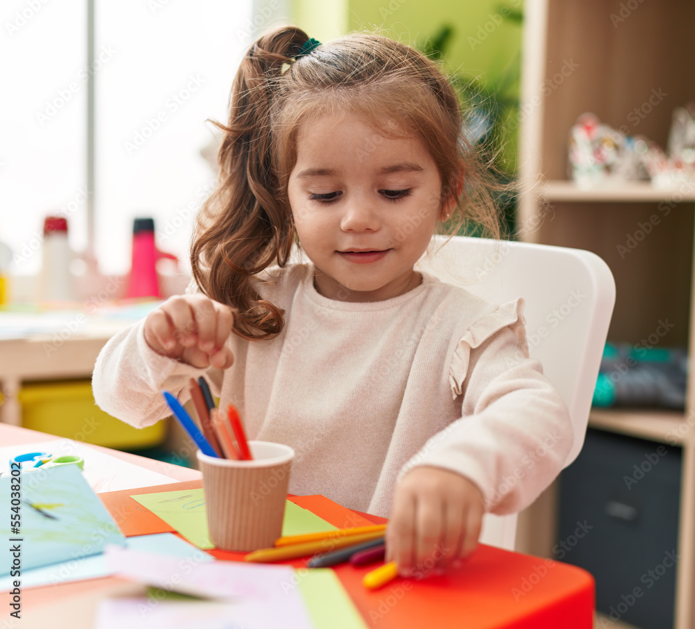 Adorable hispanic girl student sitting on table drawing on paper at kindergarten Adorable hispanic girl student sitting on table drawing on paper at kindergarten