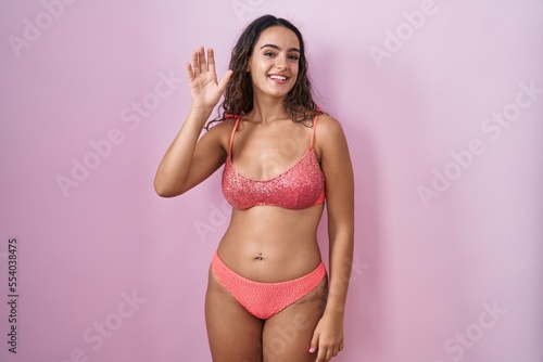 Young hispanic woman wearing lingerie over pink background waiving saying hello happy and smiling, friendly welcome gesture