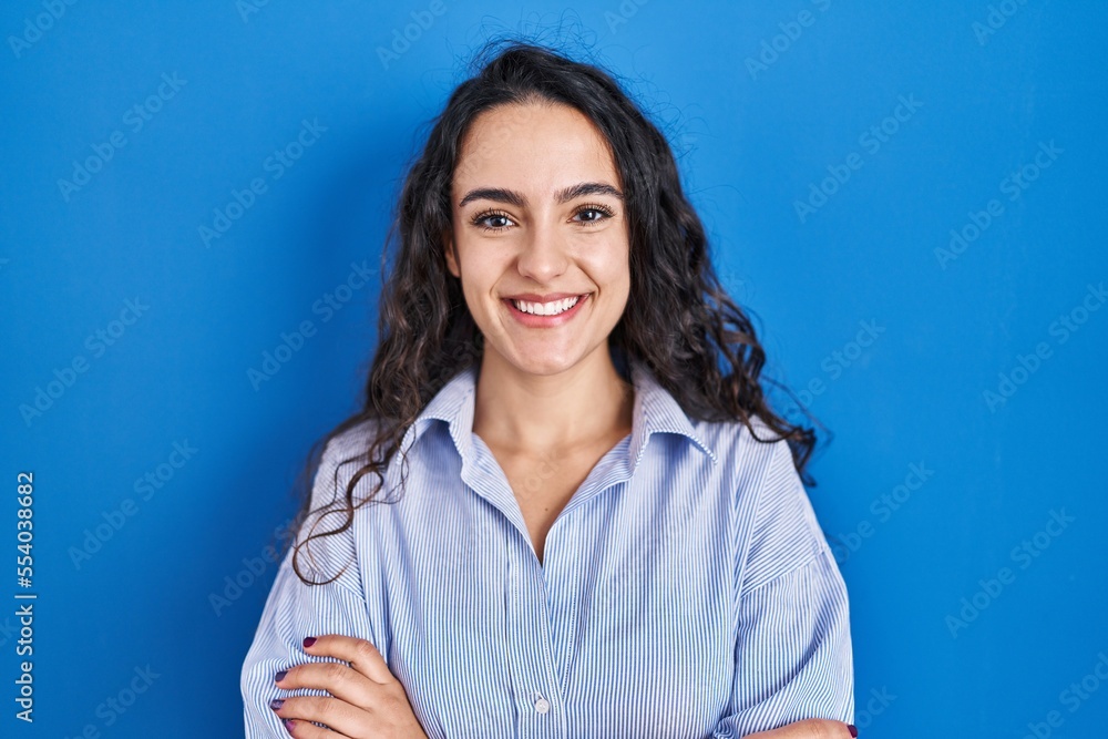 Fototapeta premium Young brunette woman standing over blue background happy face smiling with crossed arms looking at the camera. positive person.