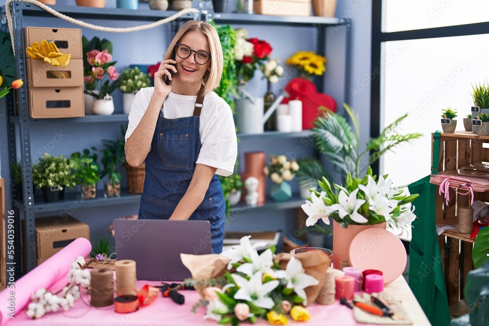 Young blonde woman florist using laptop talking on smartphone at florist shop Young blonde woman florist using laptop talking on smartphone at florist shop