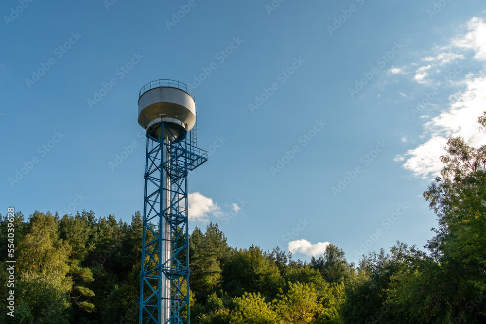 New modern water tower on the background of blue sky and forest. Methods of water storage for irrigation of agricultural crops in difficult climatic and weather conditions. A barrel with water New modern water tower on the background of blue sky and forest. Methods of water storage for irrigation of agricultural crops in difficult climatic and weather conditions. A barrel with water