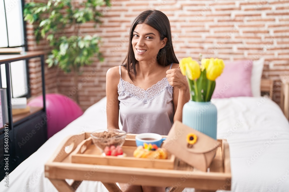 Brunette young woman eating breakfast sitting on the bed smiling happy and positive, thumb up doing excellent and approval sign