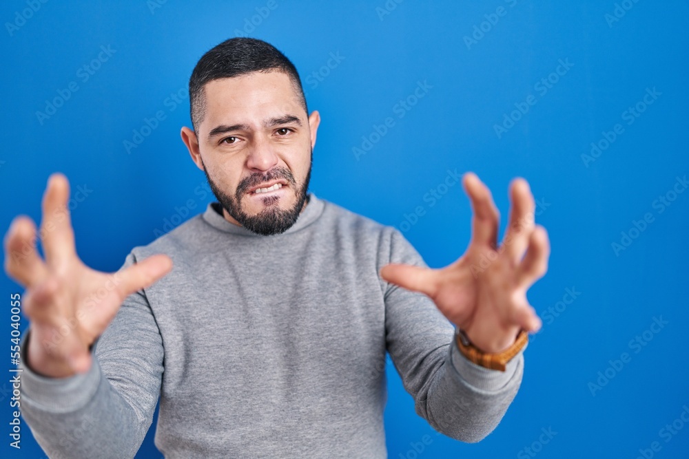 Hispanic man standing over blue background shouting frustrated with rage, hands trying to strangle, yelling mad Hispanic man standing over blue background shouting frustrated with rage, hands trying to strangle, yelling mad