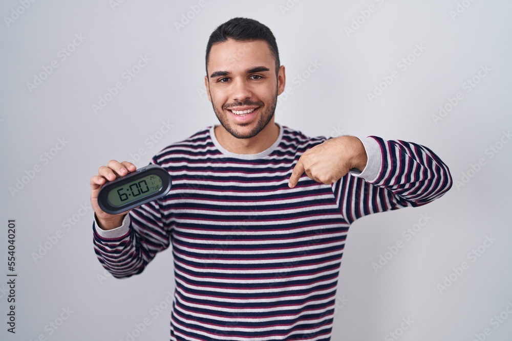 Young hispanic man wearing pyjama holding alarm clock pointing finger to one self smiling happy and proud Young hispanic man wearing pyjama holding alarm clock pointing finger to one self smiling happy and proud