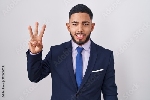 Young hispanic man wearing business suit and tie showing and pointing up with fingers number three while smiling confident and happy.