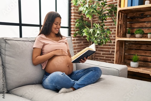 Young latin woman pregnant touching belly reading book at home