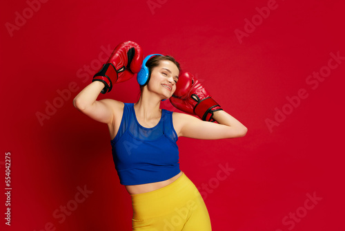 Portrait of young beautiful girl in sportive clothes posing in headphones and boxing gloves over red background