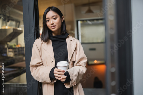 Pretty asian woman in trench coat holding coffee to go near open door of cafe.