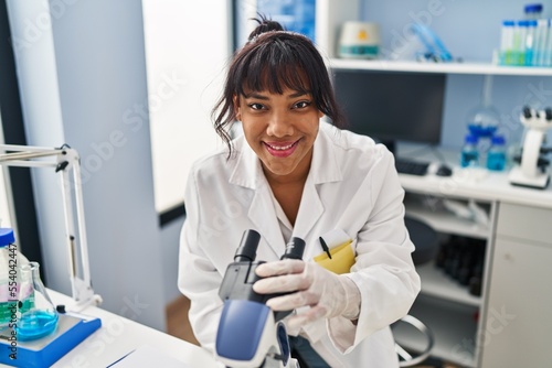 Young beautiful latin woman scientist smiling confident using microscope at laboratory