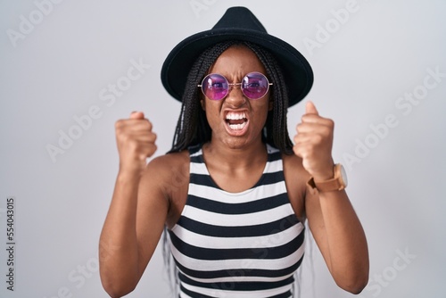 Young african american with braids wearing hat and sunglasses angry and mad raising fists frustrated and furious while shouting with anger. rage and aggressive concept.