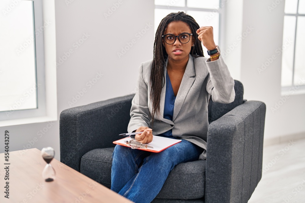 Fototapeta premium Young african american with braids working at consultation office angry and mad raising fist frustrated and furious while shouting with anger. rage and aggressive concept.