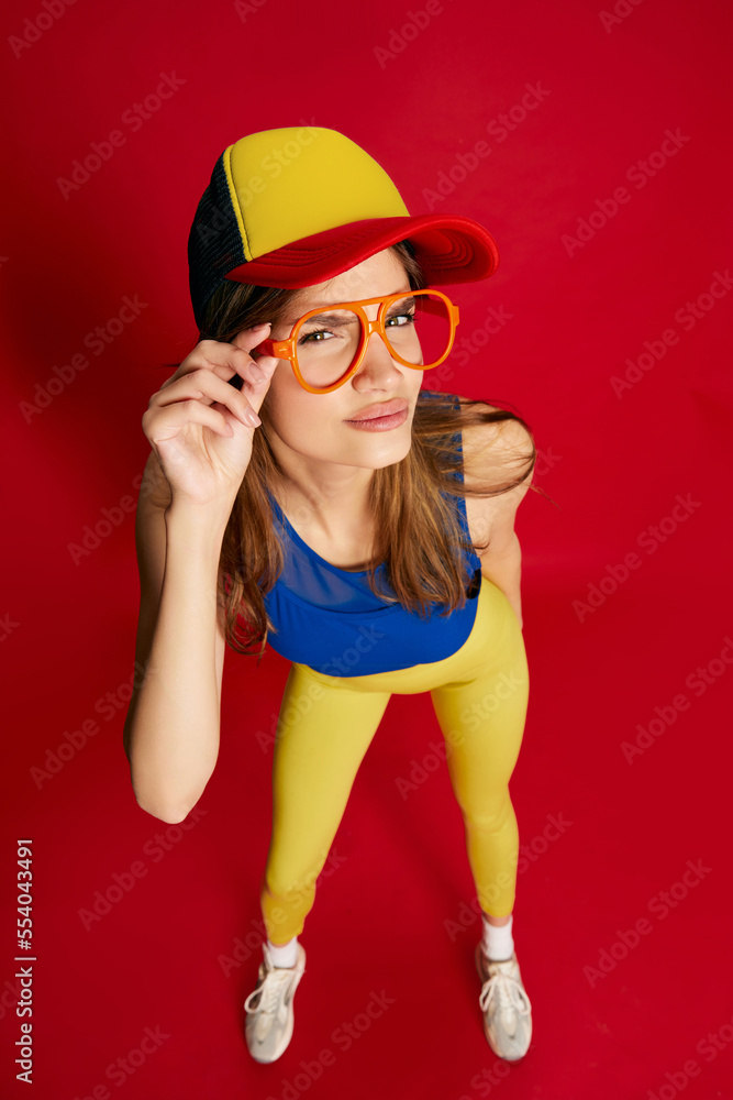 Portrait of young beautiful girl in sportive clothes, cap and glasses posing over red background. Deep look Portrait of young beautiful girl in sportive clothes, cap and glasses posing over red background. Deep look