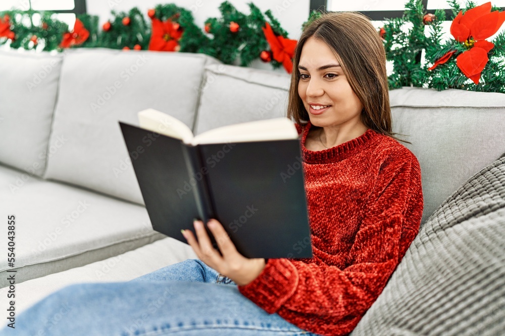 Obraz premium Young hispanic girl reading book sitting on the sofa by christmas decor at home.