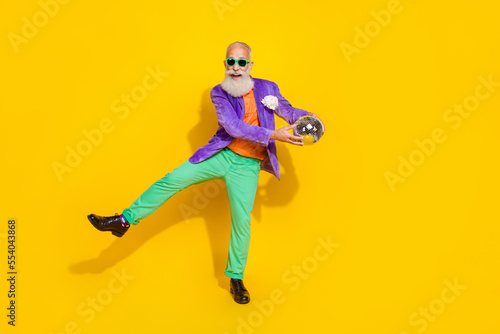 Full length photo of funny cheerful man wear purple velvet jacket dancing holding discotheque ball walking isolated yellow color background