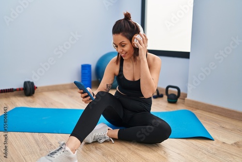Young beautiful hispanic woman sitting on yoga mat listening to music at sport center