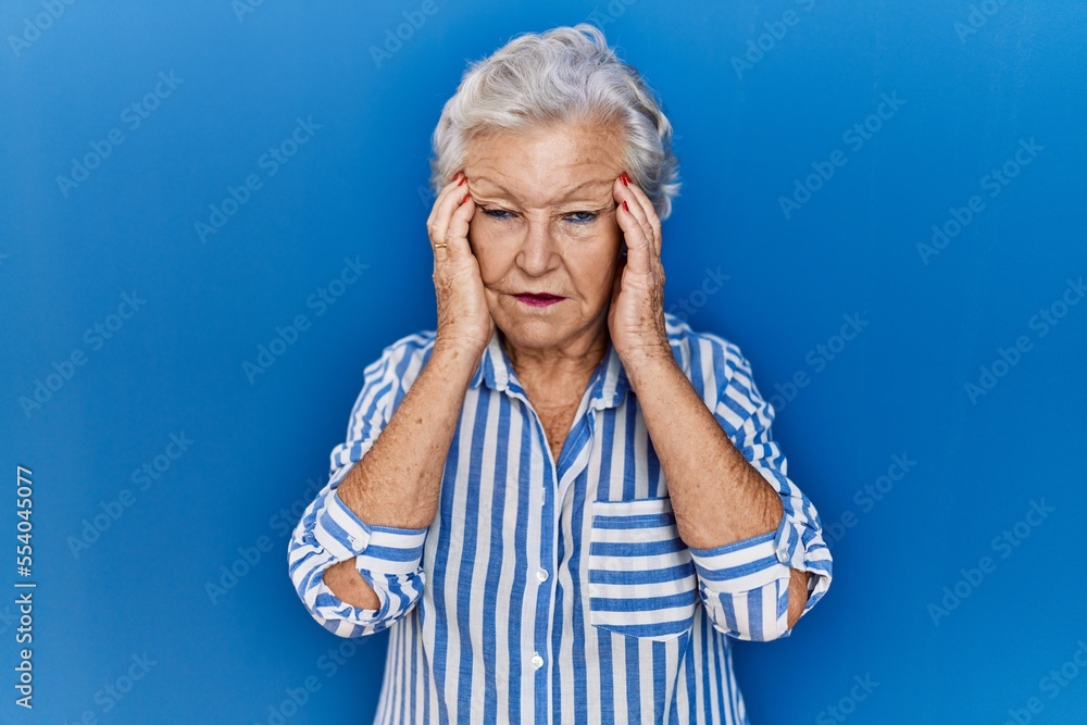 Senior woman with grey hair standing over blue background with hand on head for pain in head because stress. suffering migraine. Senior woman with grey hair standing over blue background with hand on head for pain in head because stress. suffering migraine.