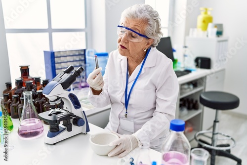 Senior grey-haired woman wearing scientist uniform holding pill at laboratory