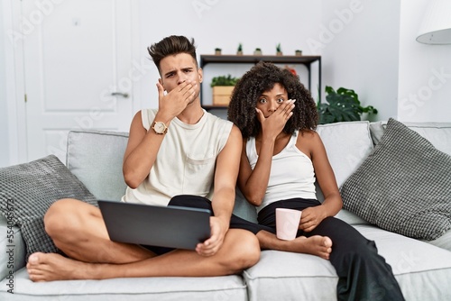 Young interracial couple using laptop at home sitting on the sofa shocked covering mouth with hands for mistake. secret concept.