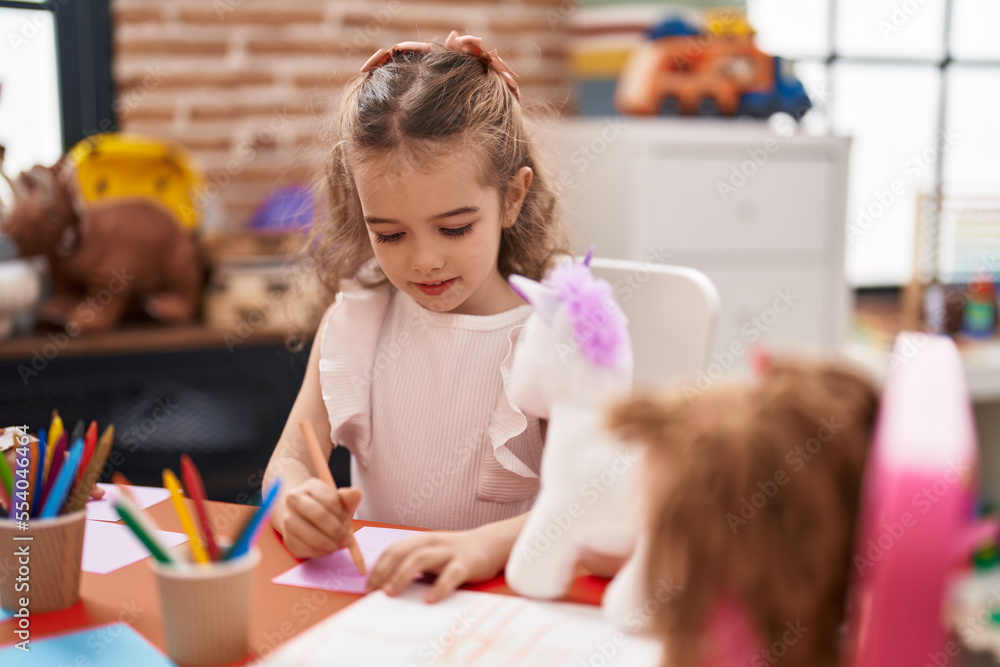 Adorable caucasian girl student sitting on table drawing on paper at classroom